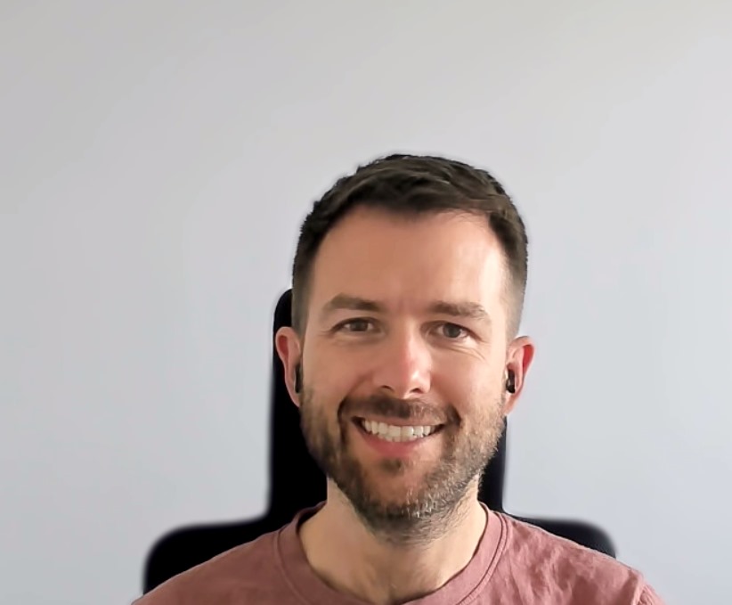 A portrait photo of a white man with brown short hair and stubble. He is looking and smiling directly at the camera, and wearing black earbuds.